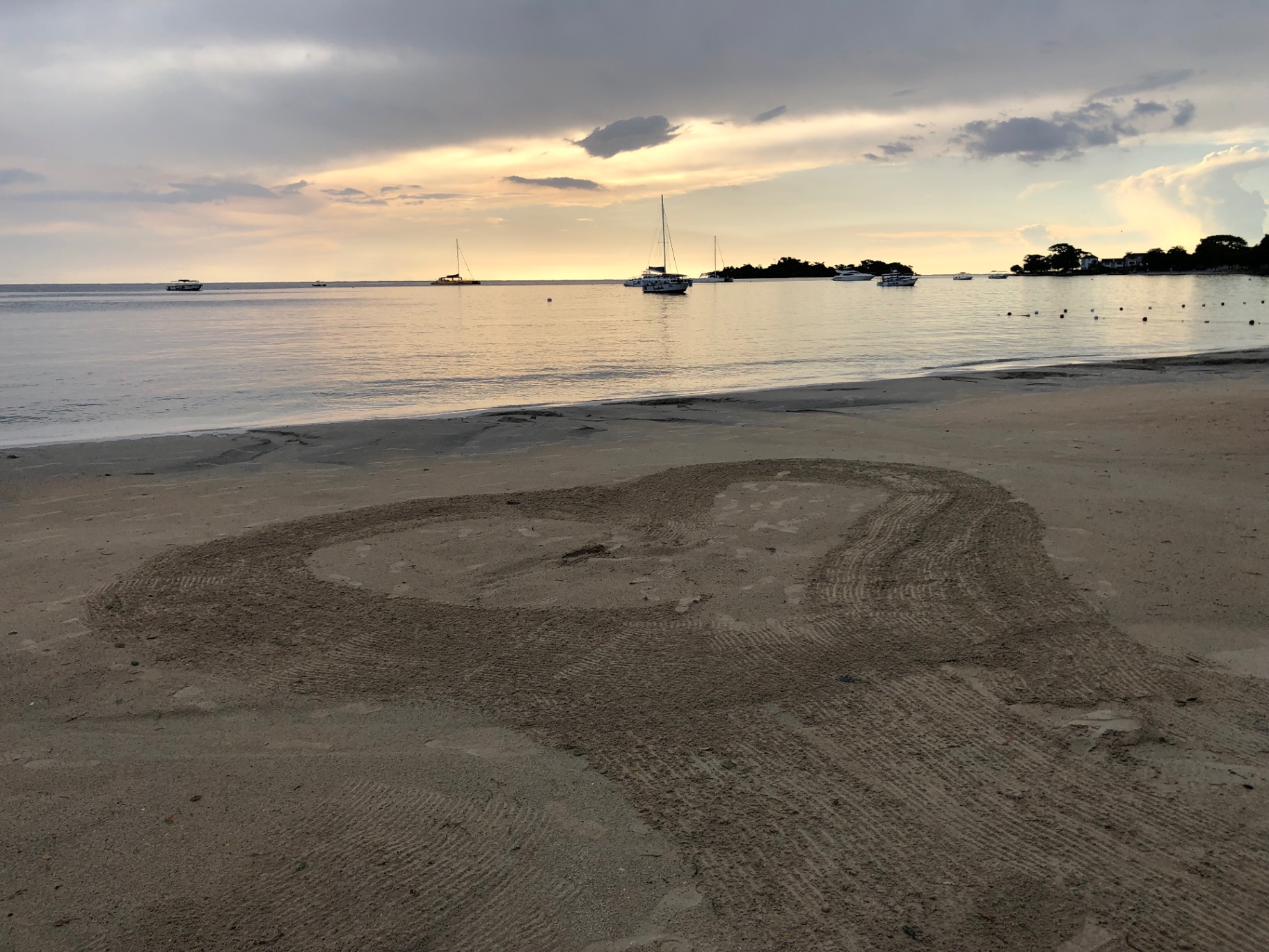 A heart raked into the sand on a quiet beach at dusk with sailboats