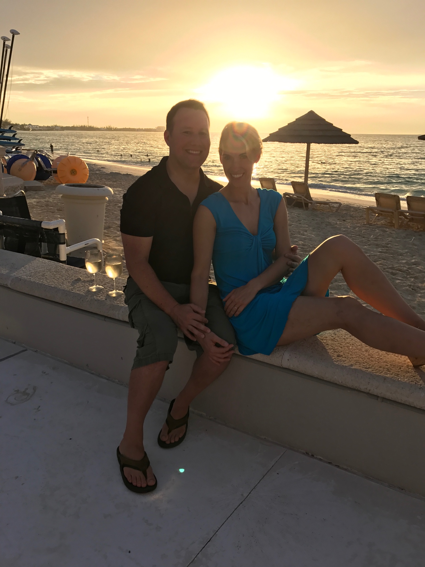 Adam and Jen Conway sitting on a beach wall at sunset with two glasses of wine