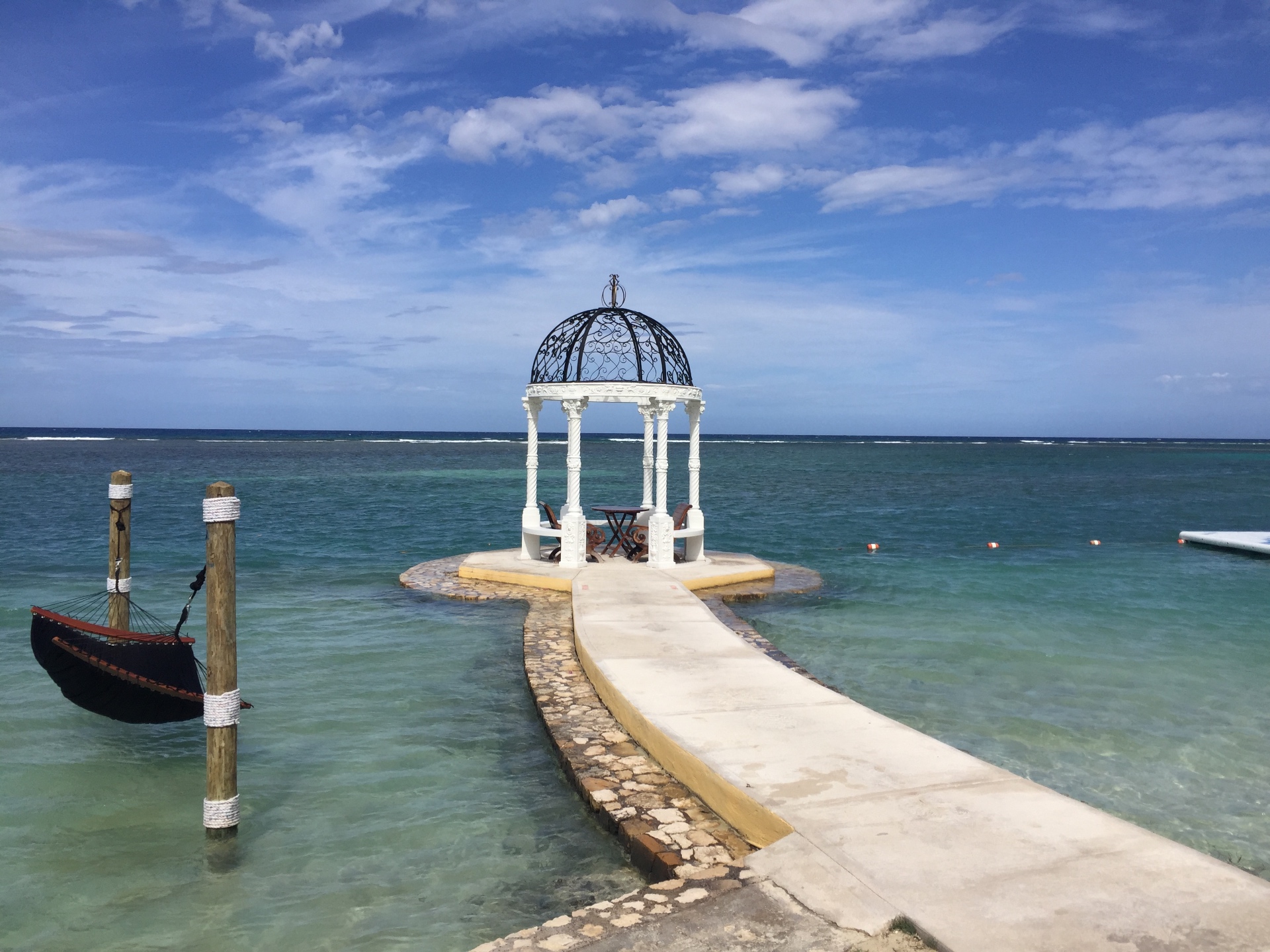 A white gazebo at the end of a stone walkway over turquoise water, with a hammock nearby