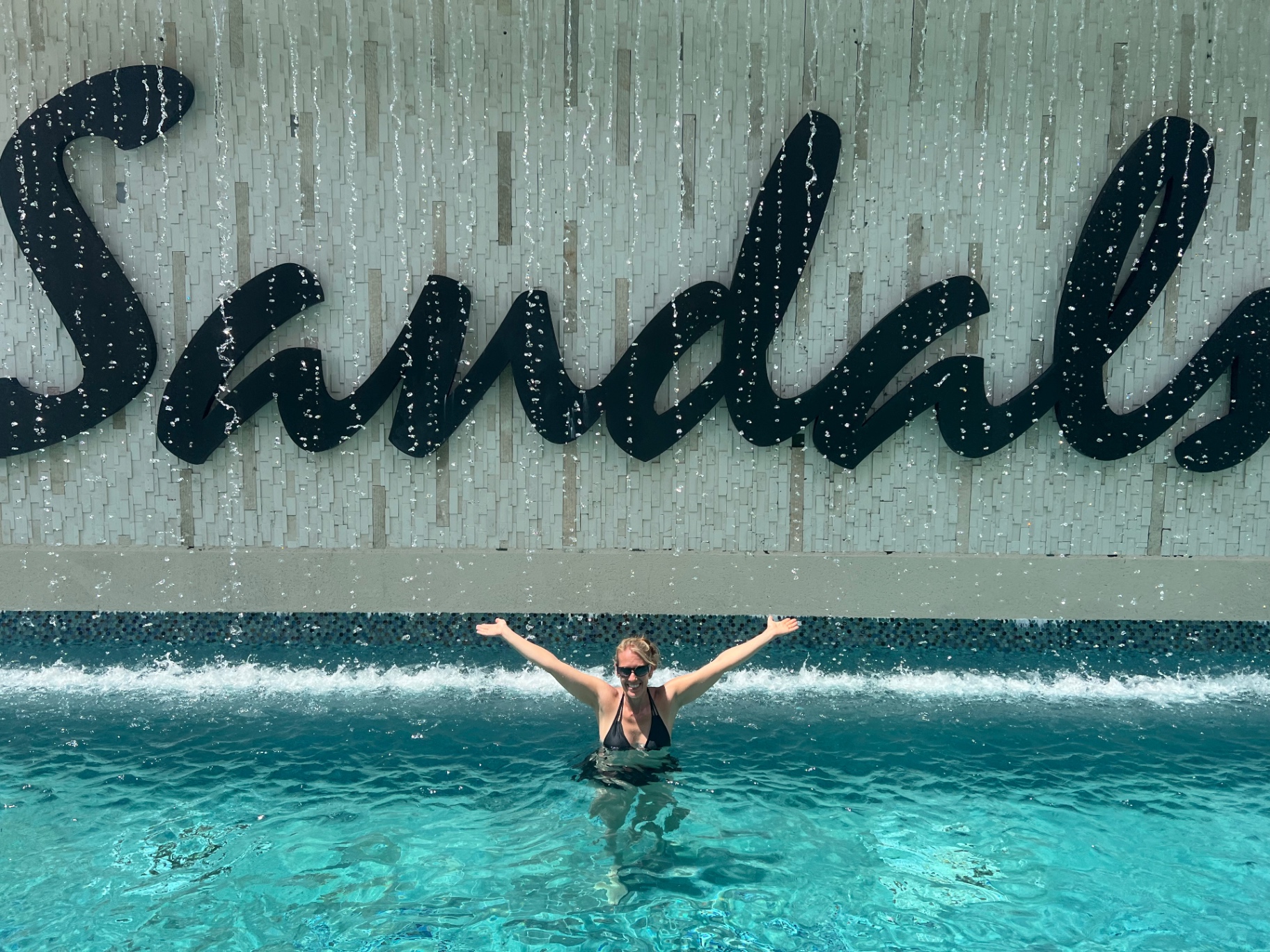 Jen Conway standing chest deep in a turquoise Sandals resort pool with arms outstretched in front of a Sandals waterfall sign