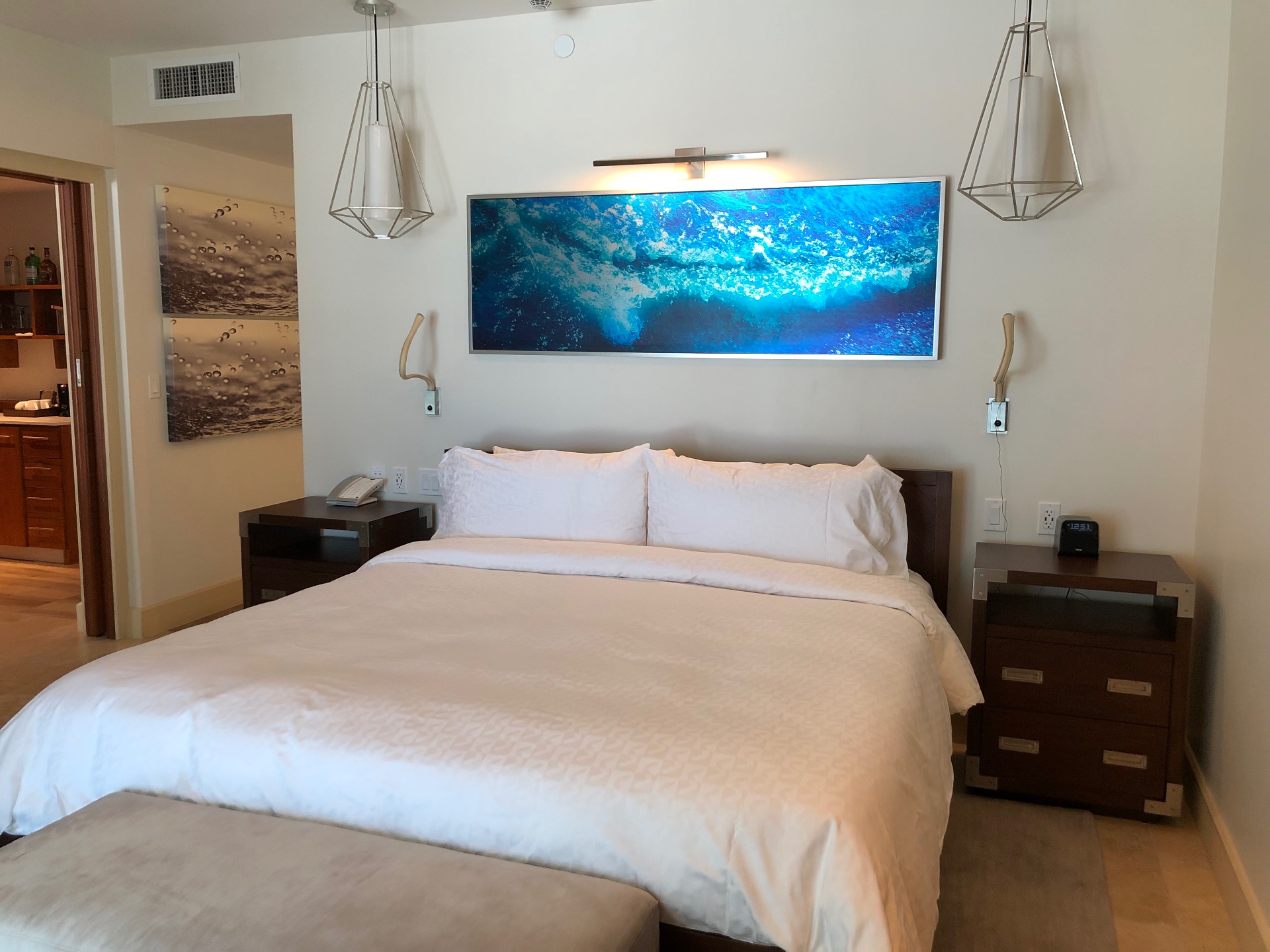 A bright resort suite bedroom with a large bed beneath a turquoise ocean artwork