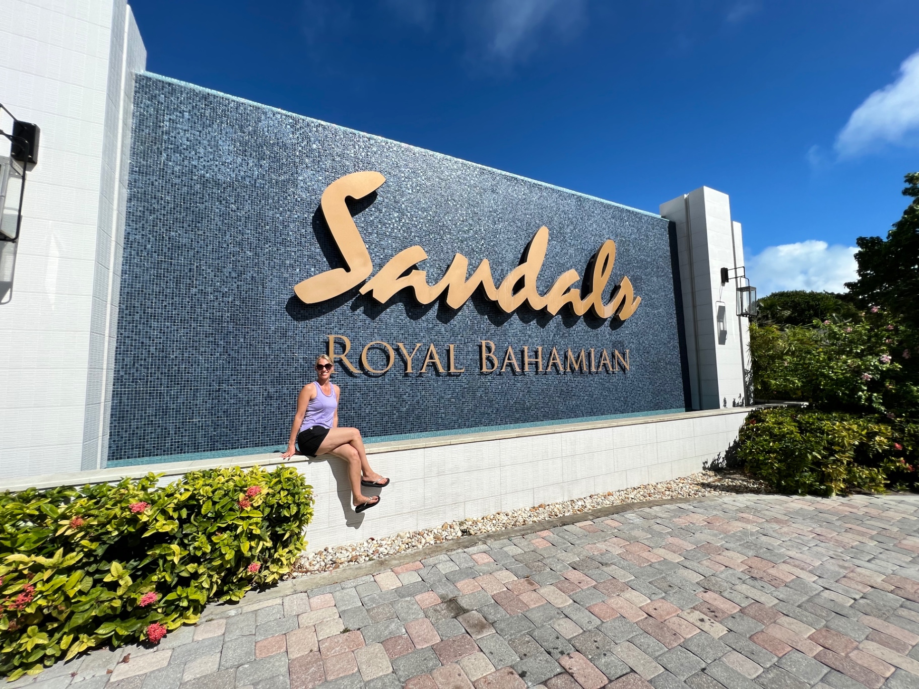 Jen seated on a ledge beneath the gold Sandals Royal Bahamian lettering on a blue mosaic wall